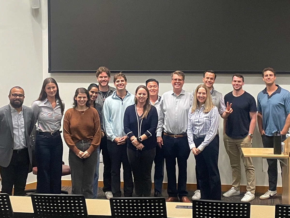 Photo: John Loper stands with a group of 12 students at the front of a classroom in front of a presentation screen, all smiling at the camera in a lecture hall setting that reflects a collaborative, educational atmosphere.