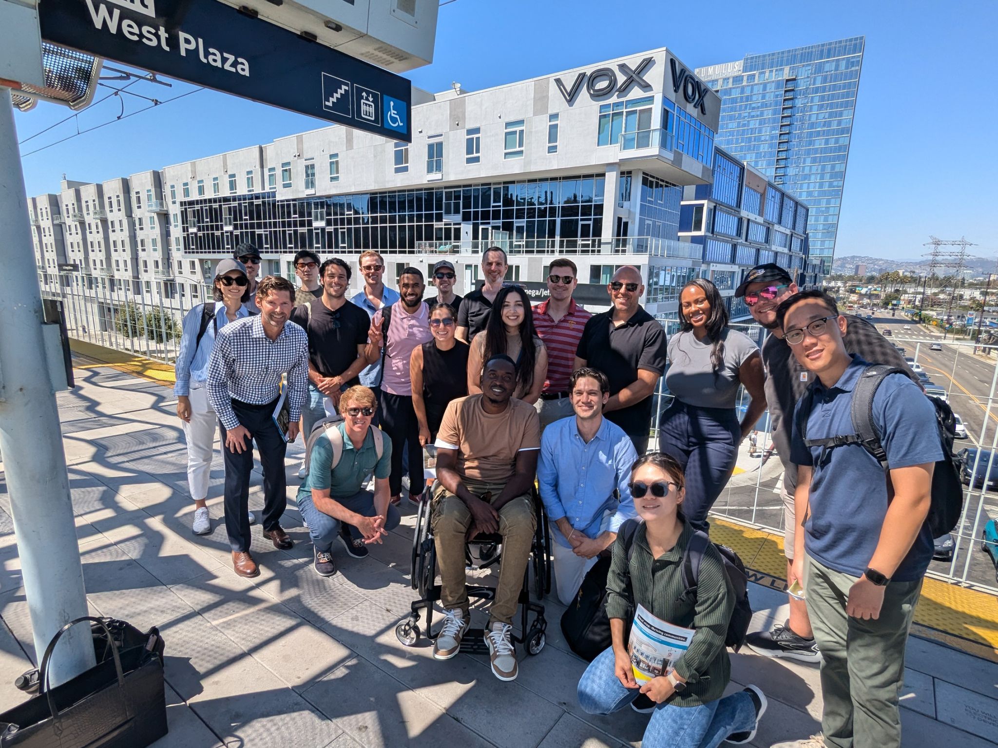 Photo: A class of students gathers on an outdoor transit platform and smiles for a group photo on a sunny day.