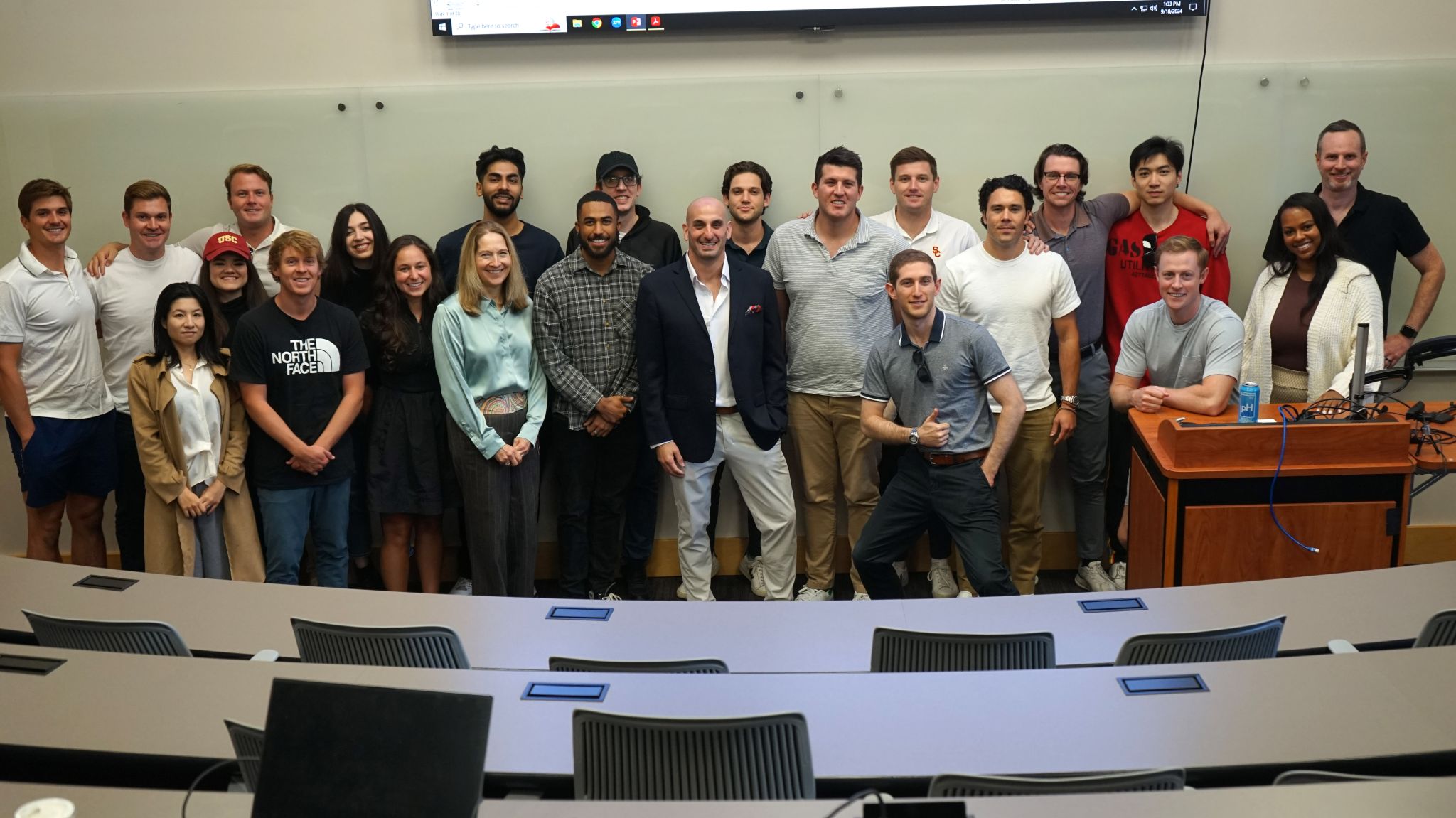 Photo: A group of about 20 students stand in a lecture hall smiling for a photo. 