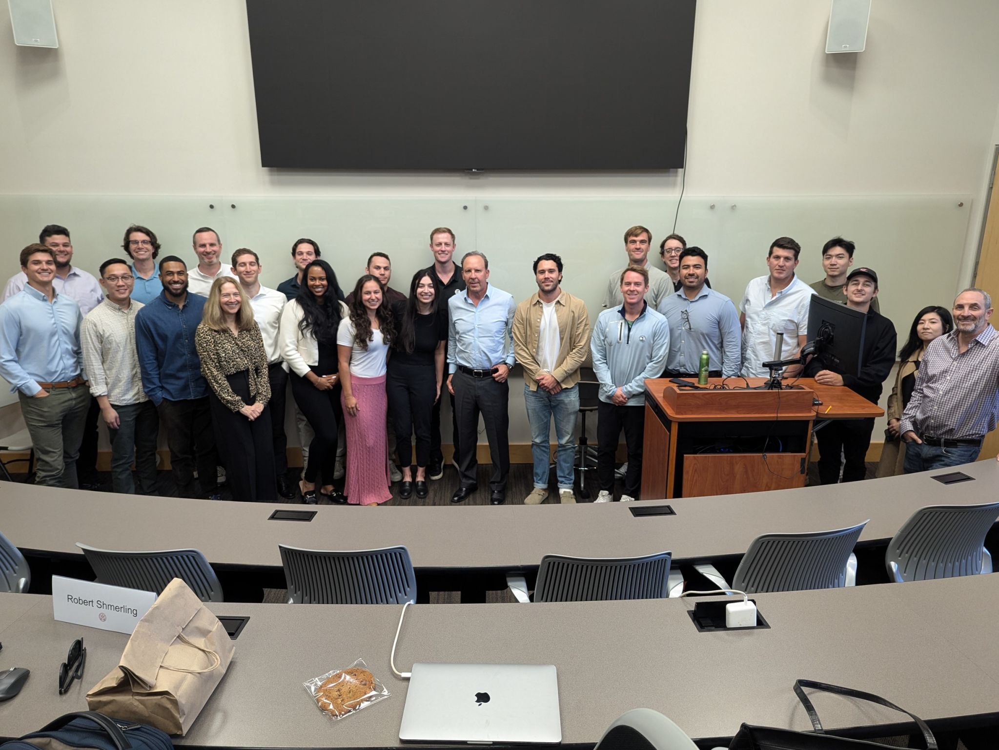 Photo: A group of about 25 students stand at the front of a lecture hall with speaker David B. Dollinger, center.